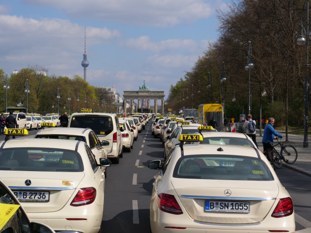 Eine lange Reihe von Taxis, die an einer belebten Straße in Berlin, Deutschland, geparkt sind, mit Fahrzeugen, Radfahrern und Fußgängern, flankiert von Laternenmasten, Bäumen und Gebäuden, einschließlich eines Bogens und eines Turms, unter einem bewölkten Himmel.