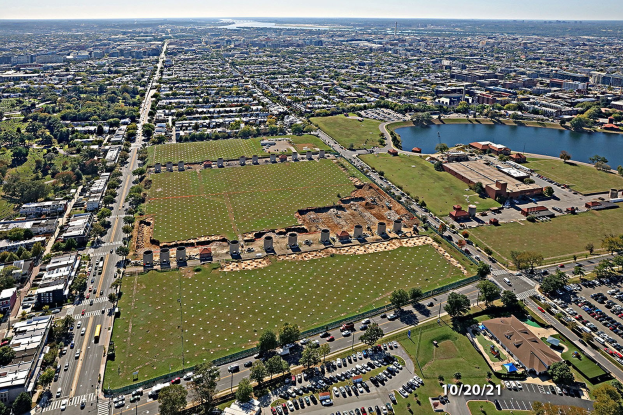 Luftaufnahme einer Stadt mit Gebäuden, Bäumen, Gras, Straßen mit Fahrzeugen, Wasser und Wolken, die einen großen zentralen Platz zeigen, der als Park geplant ist.