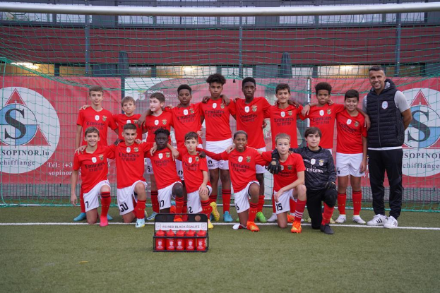 Eine Gruppe junger Männer in roten T-Shirts und schwarzen Shorts steht auf einem Fußballfeld vor einer Kiste roter Gegenstände, im Hintergrund sind Banner mit "FC Rot-Schwarz Eglaute - Champions League" zu sehen.