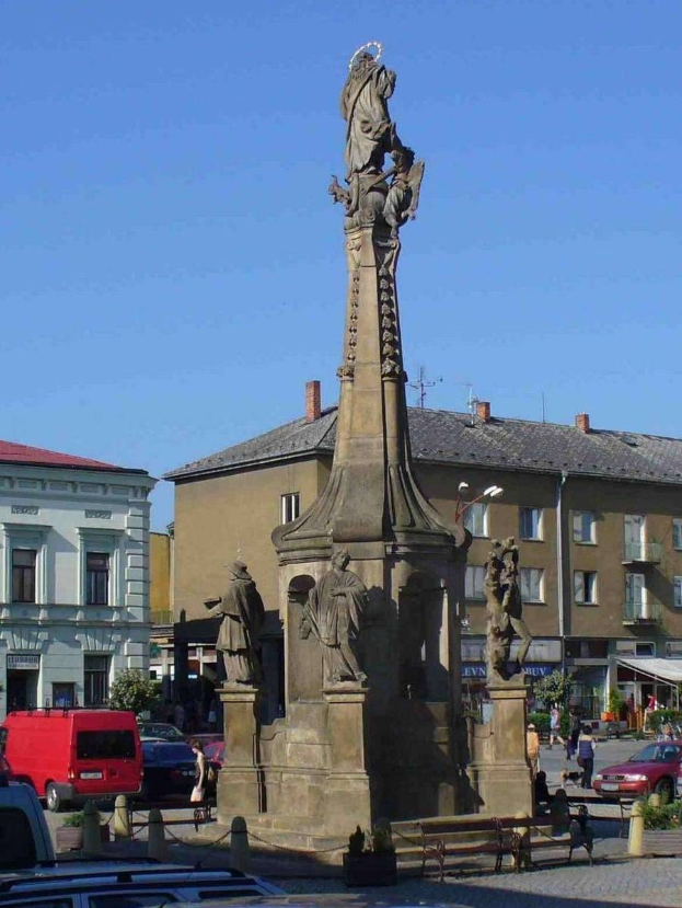 Ein Denkmal steht auf einem Platz umgeben von Gebäuden, Straßenlaternen, Verkehr, Fußgängern, Bänken, Pflanzen und einem bewölkten Himmel.
