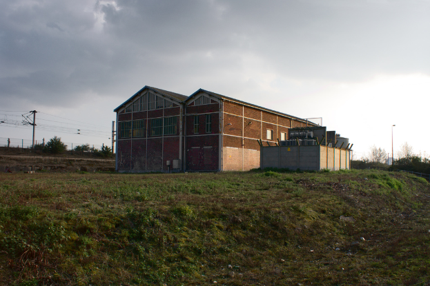 Eine Szene mit Gartenlauben, Transformatoren, Strommasten mit Kabeln, Zäunen, einer Straßenlaterne, Bäumen und einem bewölkten Himmel.
