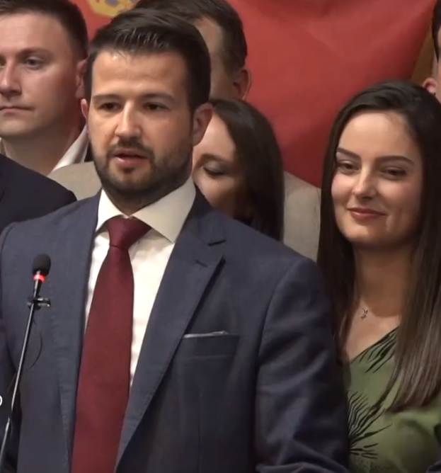 Ein Mann im Anzug und Krawatte steht an einem Rednerpult mit einem Mikrofon und einer Fahne im Hintergrund.