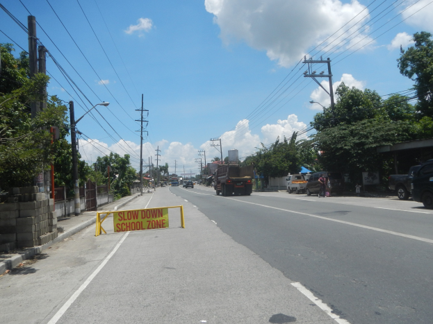 Straße mit Fahrzeugen, ein "Slow Down School Zone"-Schild, Strommasten mit Drähten, Bäume, Laternenpfähle und ein bewölkter Himmel im Hintergrund.