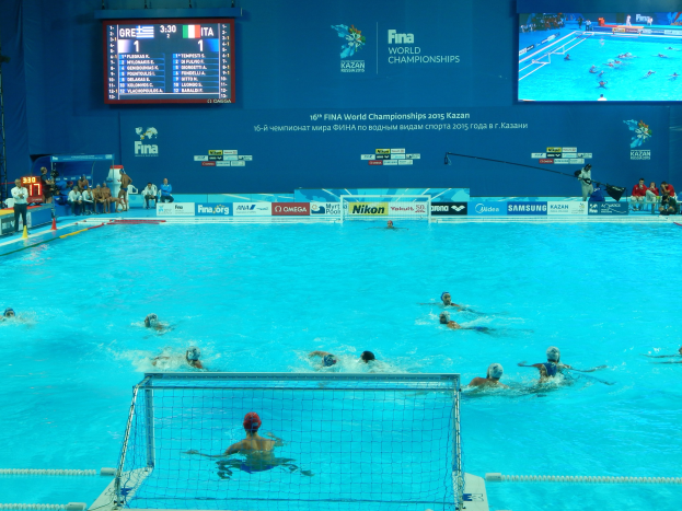 Eine Gruppe von Menschen spielt Wasserball in einem großen Pool mit einem Netz, Zuschauern auf der linken Seite und einem Bildschirm mit Text und Logos im Hintergrund bei den 2016 FINA World Championships in Kazan.