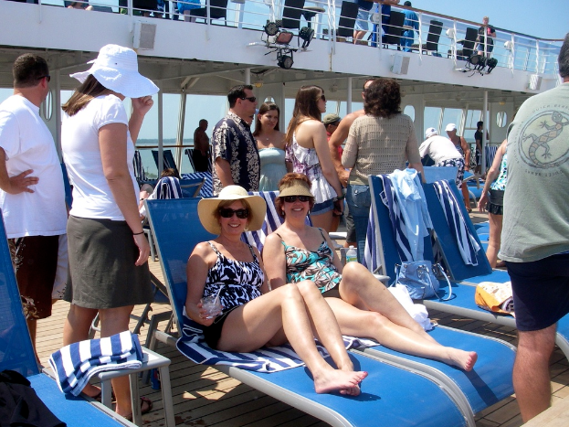 Zwei Frauen in Bikinis sitzen auf Stühlen mit vielen Menschen drumherum unter einem Himmel.