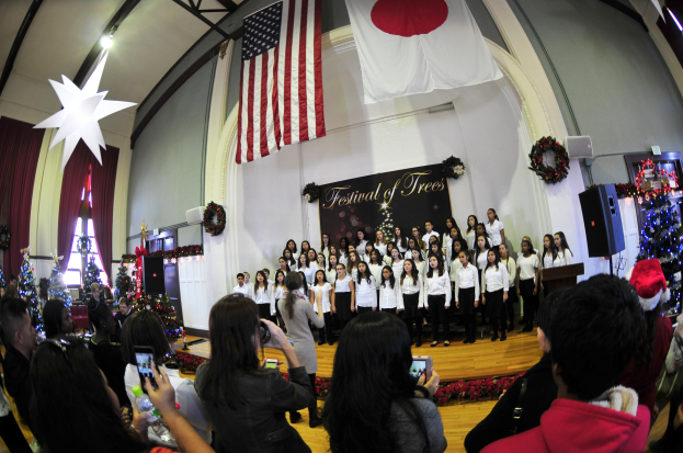 Gruppe von Menschen auf einer Bühne mit Weihnachtsdekoration, einem Rednerpult, Lautsprechern, einer Tafel mit Text, Vorhängen, Flaggen und einer Menge darunter mit Kameras und Handys.