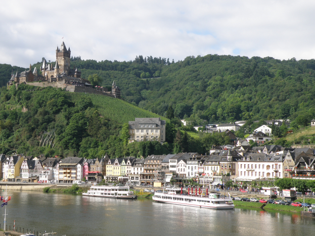 Ein malerischer Blick auf den Rhein in Deutschland, mit einer Burg auf einem Hügel im Hintergrund, Booten auf dem Fluss, Fahrzeugen auf einer nahen Straße und einer bewölkten Himmel.