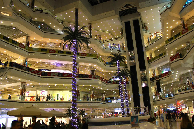 Ein Innenblick auf ein Einkaufszentrum mit Trees, Schirmen mit St√∂cken, Personenaufz√ľgen, Namensschildern, St√§nden, Lichtern, Leuchttafeln und einer Gruppe von Menschen.