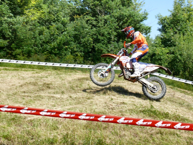 Eine Person mit Helm fährt ein Fahrrad, umgeben von Absperrband, mit Bäumen, Gras und Himmel im Hintergrund.