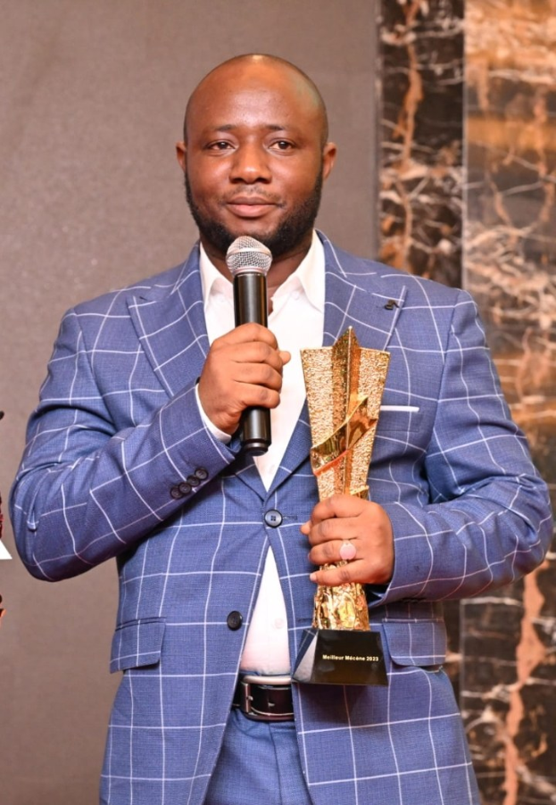 Ein Mann in einem blauen Anzug hält ein Mikrofon und eine Trophäe und strahlt selbstbewusst, als würde er einen Sieg feiern.