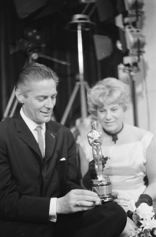 Ein Mann in einem schwarzen Blazer, weißem Hemd und Krawatte hält eine Oscar-Statue und sitzt neben einer Frau, die einen Blumenstrauß hält, mit Lichtern im Hintergrund, alles in Schwarz-Weiß.