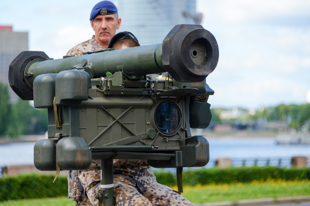 Zwei Personen in militärischer Uniform mit grünen Waffen vor einem See mit Gebäuden, Bäumen und einem klaren Himmel.