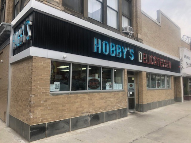 Außenansicht von Hobby's Delicatessen in Chicago, Illinois, mit Glasfenstern und -türen, einem Namensschild, einer Laterne davor und einem bewölkten Himmel im Hintergrund.