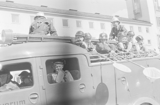 Schwarzes und weißes Foto von Kindern mit Helmen, die im Rücken eines Feuerwagens sitzen, mit einem Fenster aufweisenden Gebäude im Hintergrund.