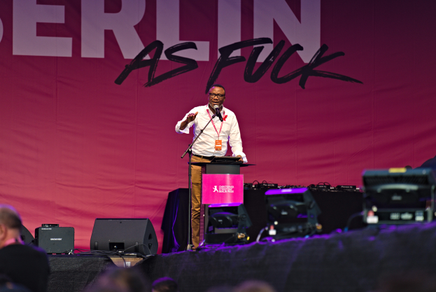 Ein Mann in einem weißen Hemd und braunen Hosen steht auf einer Bühne und hält ein Mikrofon in der Hand, hinter ihm ein pinker Banner mit der Aufschrift "Berlin as Fuck" und Lautsprecher auf der Bühne.