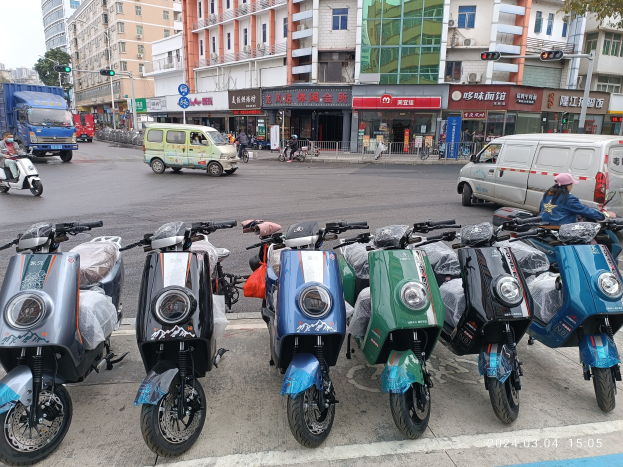 Eine Reihe von E-Scootern, die auf einer Straße geparkt sind, mit einer Person in einer Mütze davor, vor einem Hintergrund aus Gebäuden, Ampeln, Bäumen und einem klaren blauen Himmel.