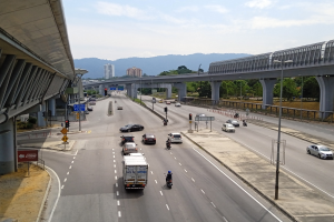 Eine vielbefahrene Autobahn mit starkem Verkehr neben einer Brücke, mit Straßenlaternen, Verkehrsampeln, Schildern, Bäumen, Gebäuden, Hügeln und einem bewölkten Himmel im Hintergrund.