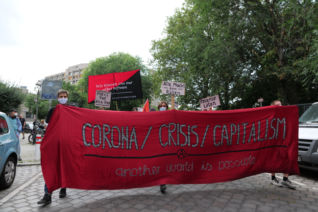 Eine Gruppe von Menschen marschiert mit einer roten Fahne, auf der 'Corona-Krise/Kapitalismus' steht, eine Straße entlang. Parkende Fahrzeuge stehen auf beiden Seiten und im Hintergrund sind Gebäude, Bäume und ein klarer blauer Himmel zu sehen.