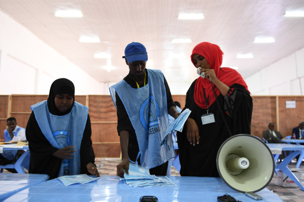 Eine Gruppe von Menschen steht um einen Tisch mit einem Megafon und Papieren herum, was wie eine Wahllokalität aussieht, mit anderen im Hintergrund unter Deckenlampen sitzend und stehend.