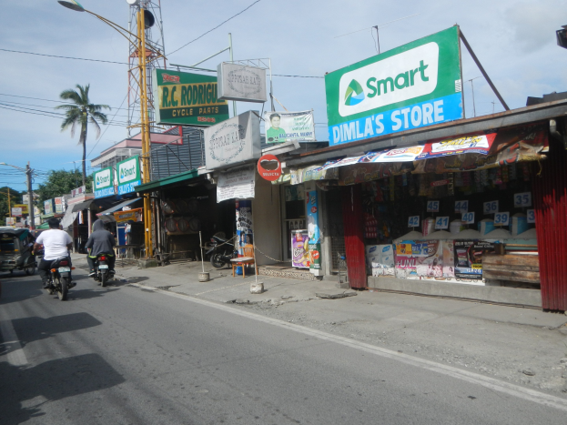 Eine belebte Straße mit Motorrädern, Autos, Gebäuden, Straßenschildern, Strommasten, Drähte, Straßenlaternen, Bäume und einem bewölkten Himmel.