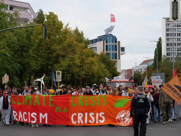 Eine Gruppe von Menschen marschiert eine von Bäumen gesäumte Straße entlang und hält ein Banner mit der Aufschrift "Klima-Krise ist eine Krise", mit Gebäuden und einem klaren blauen Himmel im Hintergrund.