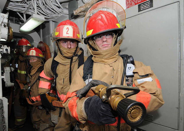 Gruppe von Feuerwehrleuten in Uniform in einem Raum mit Feuerlöscher, einer Tafel mit Text auf der rechten Seite und Hintergrundgegenständen wie Kabeln und einer Lampe.