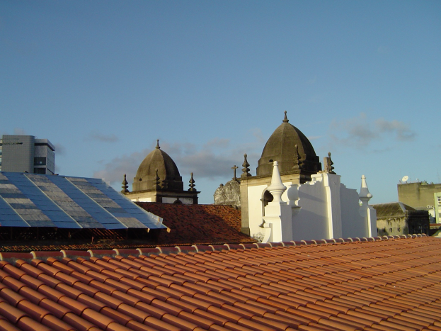 Stadtpanorama mit Gebäuden im Vordergrund und einem blauen Himmel im Hintergrund, das Solarpanels auf einem Dach zeigt, was den Einsatz erneuerbarer Energie anzeigt.