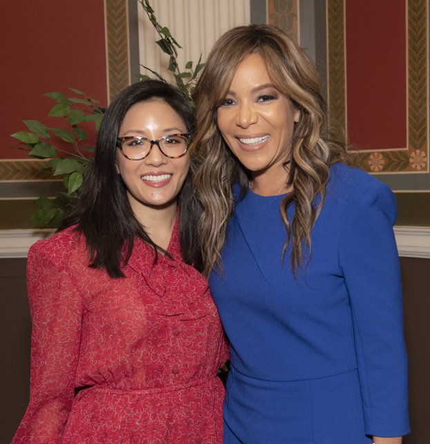 Zwei Frauen, die nebeneinander auf einem Event lächeln, eine in einem blauen Kleid und die andere in einem roten Kleid mit Brille, vor einem Hintergrund aus Pflanzen und Vorhängen.