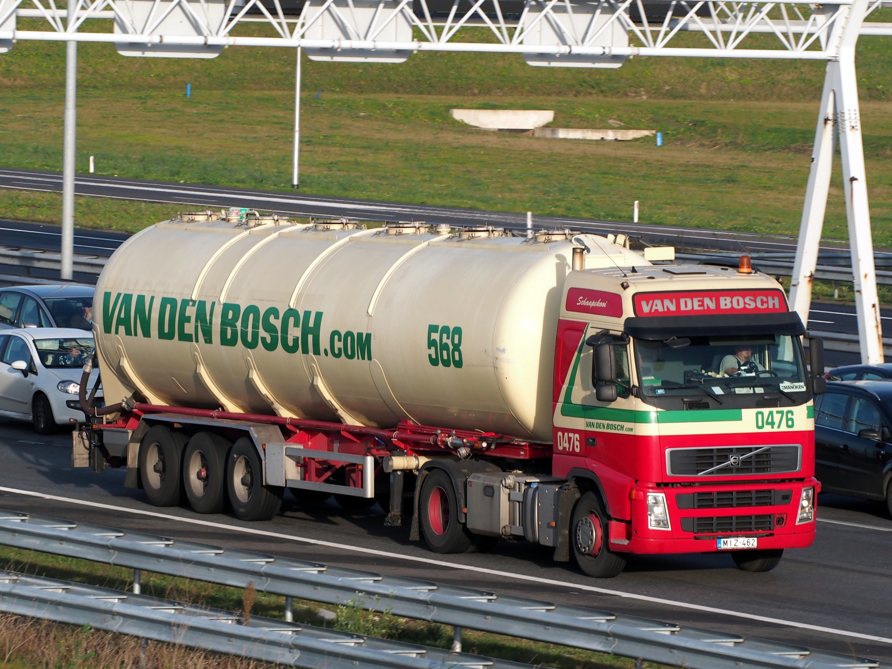 Ein großer weißer, roter und grüner Tanklastwagen fährt auf einer Autobahn, umgeben von anderen Fahrzeugen, mit einer Leitplanke am Straßenrand, Gras im Hintergrund und einem Eisenrahmenbauwerk.