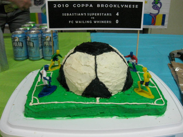 Ein kuchen in form eines fussballstadions mit einem fussball und vier spielern, umgeben von softdrink-dosen, einem banner im hintergrund und einer person oben links.