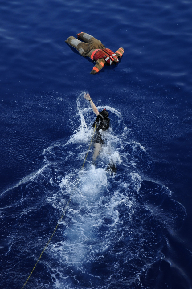 Ein Retter in einem Rettungswesten, der an einem Seil über ruhigem, klarem Wasser hängt, bei strahlendem Sonnenschein.