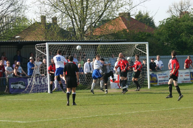 Spieler spielen Fußball auf einem Feld mit einem Tornetz und Zuschauern dahinter, mit Bäumen und Häusern im Hintergrund.