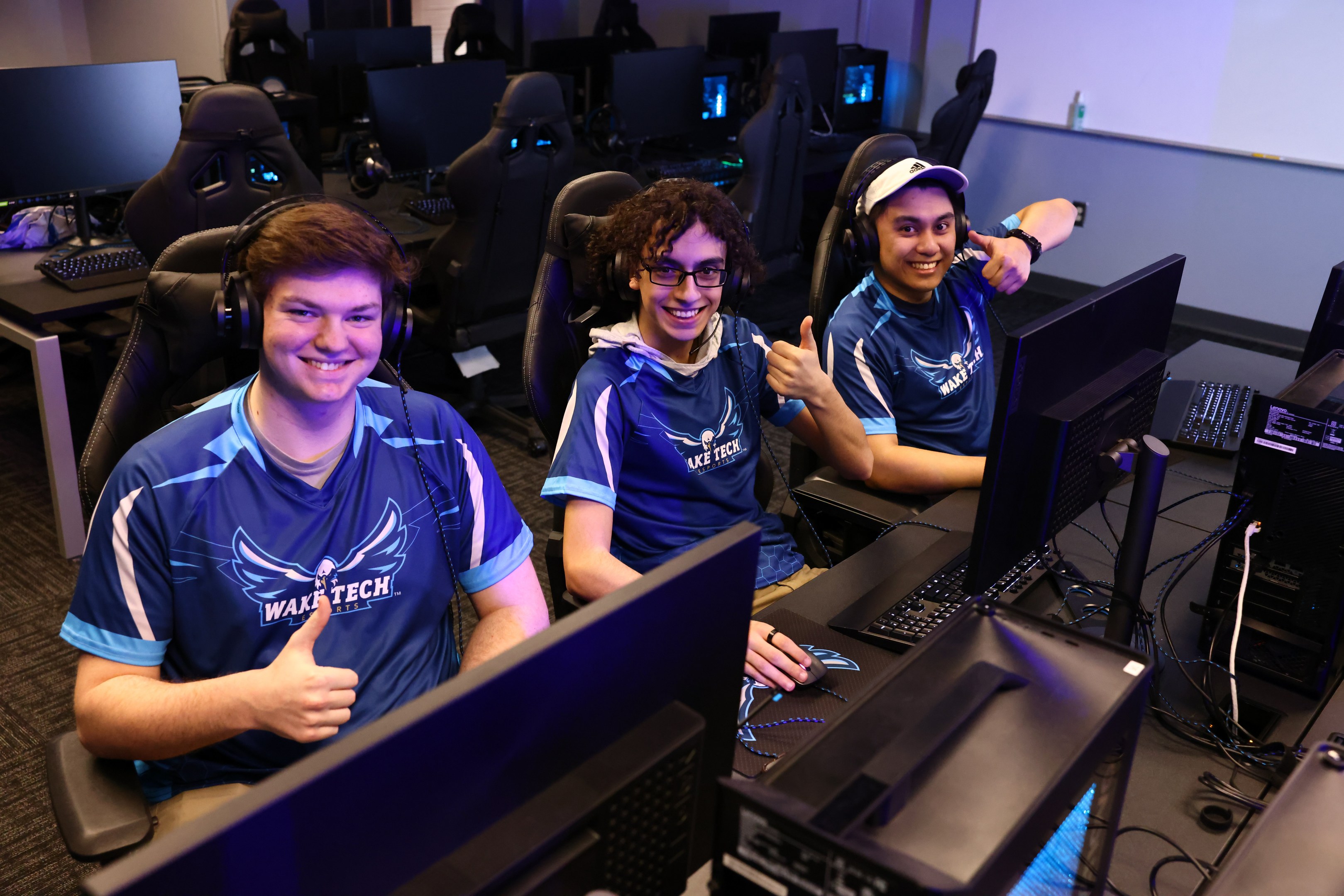 Drei junge Männer in Headsets lächeln, während sie bei einer E-Sport-Gaming-Sitzung an Computern sitzen, mit Monitoren, Tastaturen und Kabeln auf dem Schreibtisch.