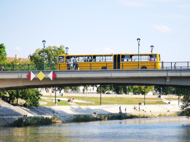 Ein gelber Schulbus fährt über eine Brücke über einen Fluss, mit Menschen, die über die Brücke gehen und stehen, umgeben von saftigem Grün und Bäumen, mit Gebäuden und einem klaren blauen Himmel im Hintergrund.