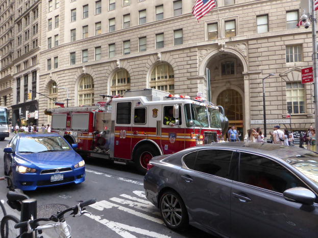 Ein roter Feuerwehrauto auf einer Straße in New York City geparkt mit Fahrzeugen, Fußgängern, Fahrrädern, Gebäuden, Laternen und Fahnenmasten im Hintergrund.