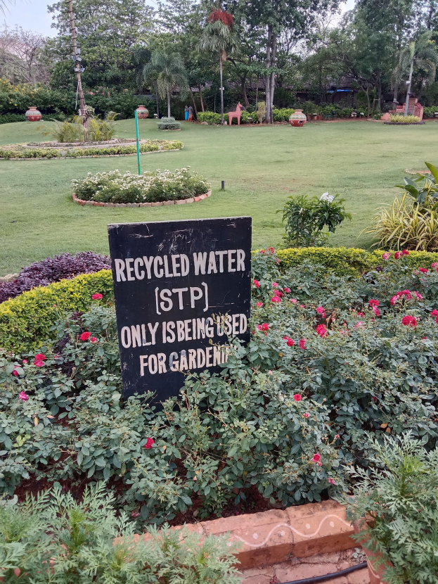 Ein Garten mit einem Schild, auf dem "Recycled Water (STP) Only Used for Gardening" steht, umgeben von grünem Gras, bunten Blumen, hohen Bäumen und einem klaren blauen Himmel.