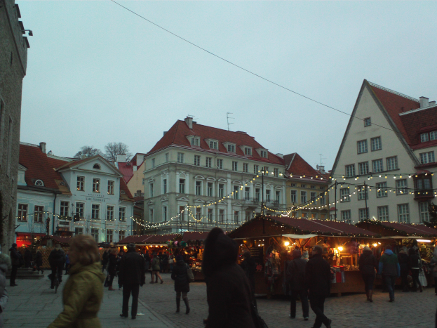 Ein lebendiger Weihnachtsmarkt in Tallinn, Estland, mit Menschen, die spazieren gehen, beleuchteten Ständen, Gebäuden, Bäumen und einem klaren blauen Himmel im Hintergrund und Text am unteren Rand des Bildes.