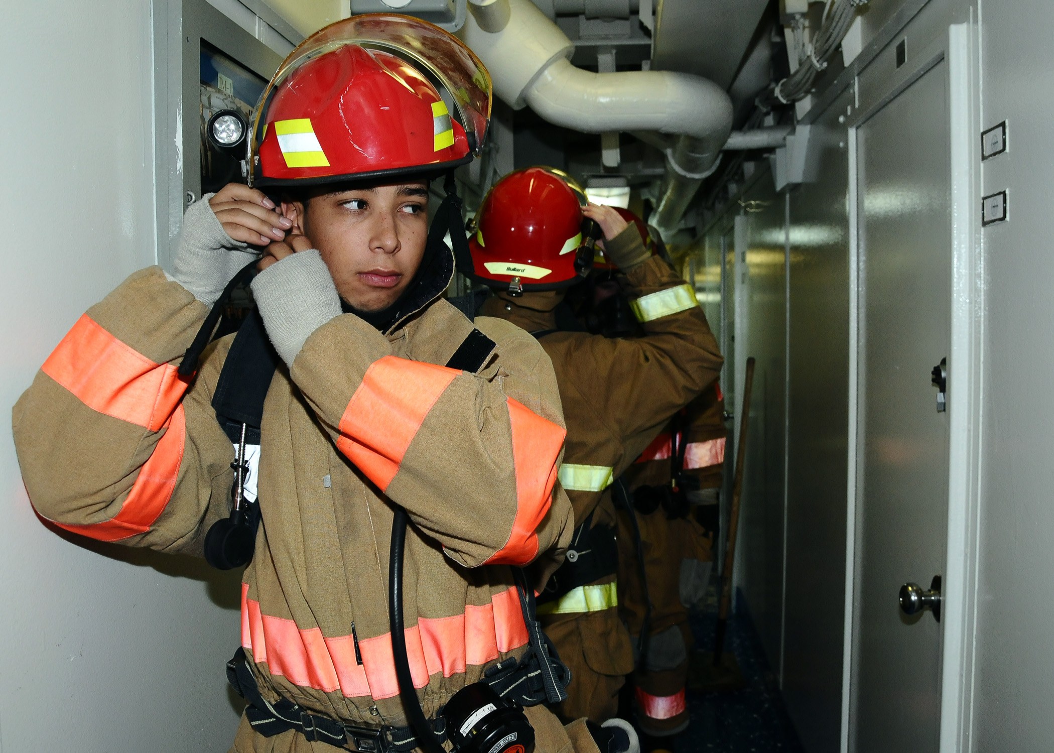 Feuerwehr in Uniform, die zusammen in einem Raum mit einer Tür, Rohren und Übungsgeräten im Hintergrund steht.