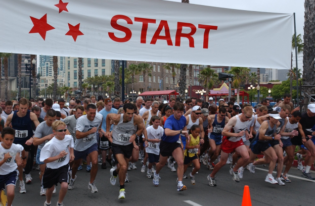 Gruppe von Läufern bei einem Marathon, die an einer Verkehrskegel vorbei läuft, mit einem Banner und Gebäuden im Hintergrund unter einem klaren blauen Himmel.